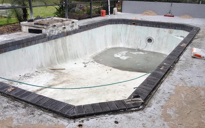 Empty swimming pool undergoing resurfacing with exposed concrete, surrounded by tools and materials, for Kristi's Pool Ser...