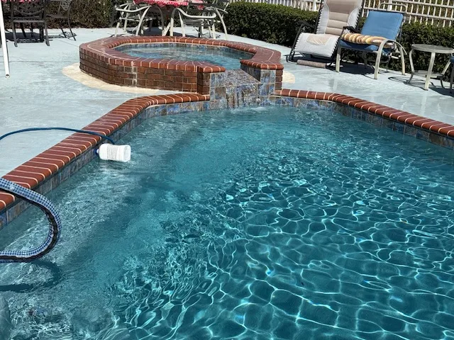 Clear blue pool with brick-lined hot tub, surrounded by patio furniture, serviced by Kristi's Pool Service.